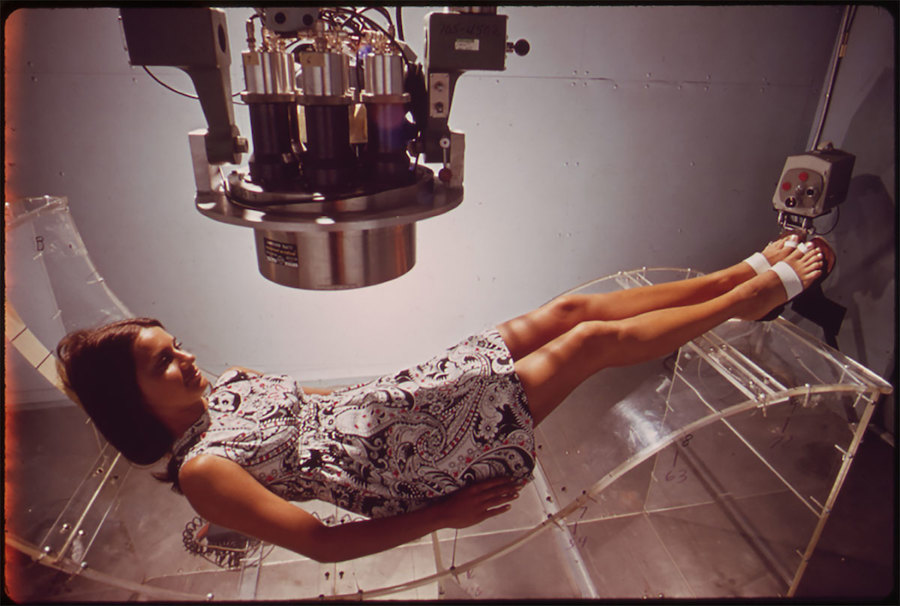 A chair-sized metal cylinder with intricate wiring is suspended from the ceiling over a curved bed where a woman is lying.