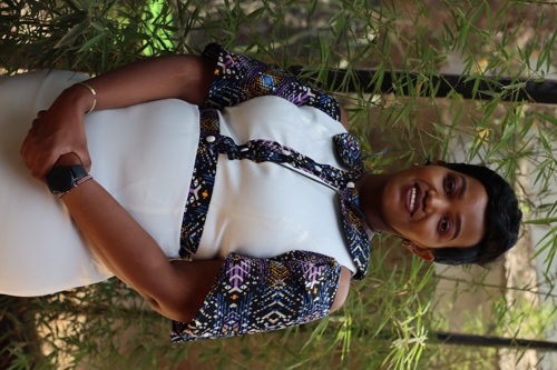 Dr. Shikoh Gitau stands in front of bamboo trees.  She is African with short black hair, a big smile, and a white dress.